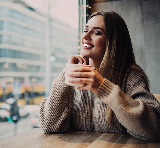 Woman in brown sweater holding mug smiling looking out window